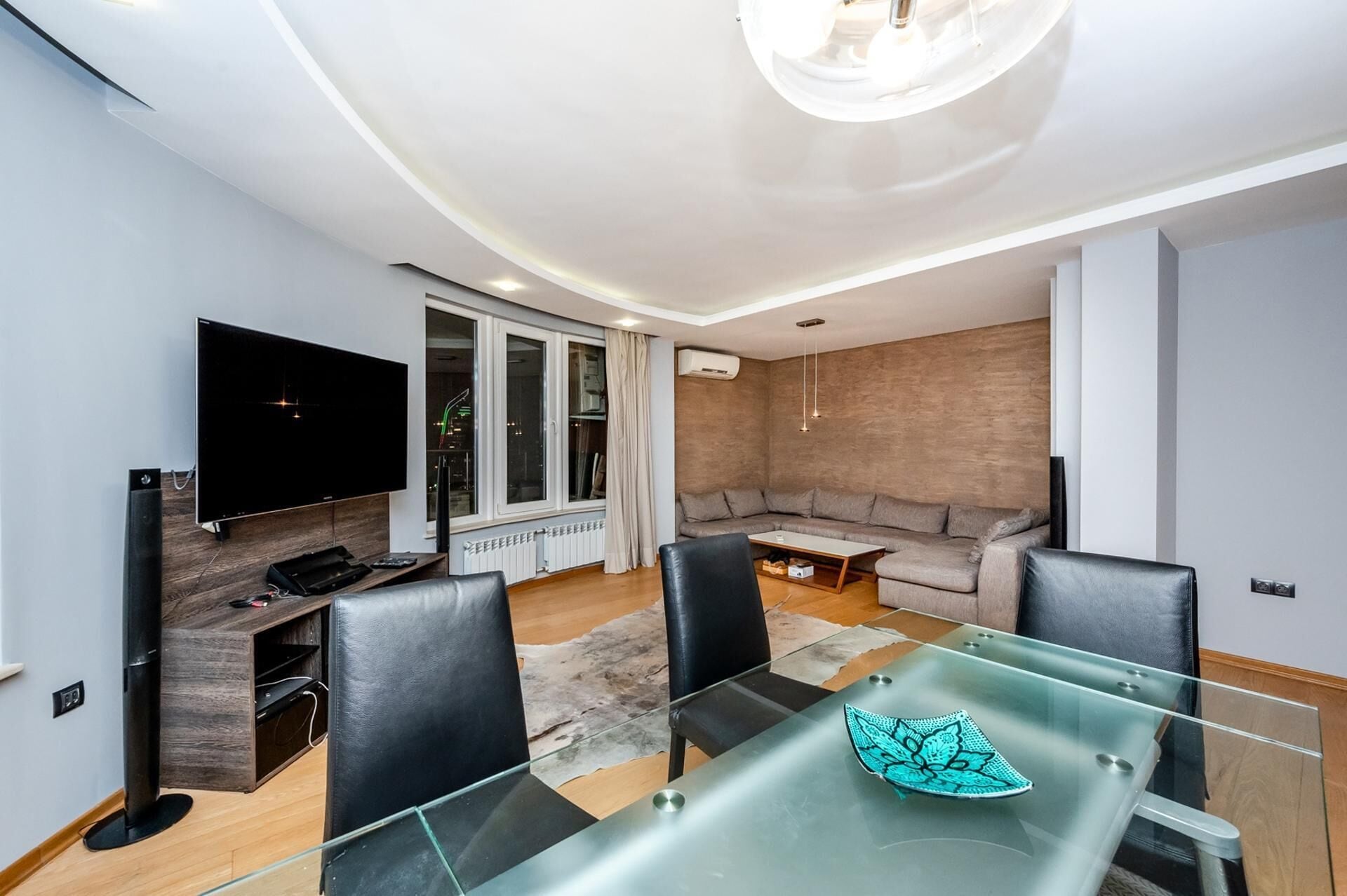 Un salón moderno con televisor de pantalla plana, una mesa de comedor de vidrio, sillas de cuero, un sofá modular y un gran ventanal con cortinas.
