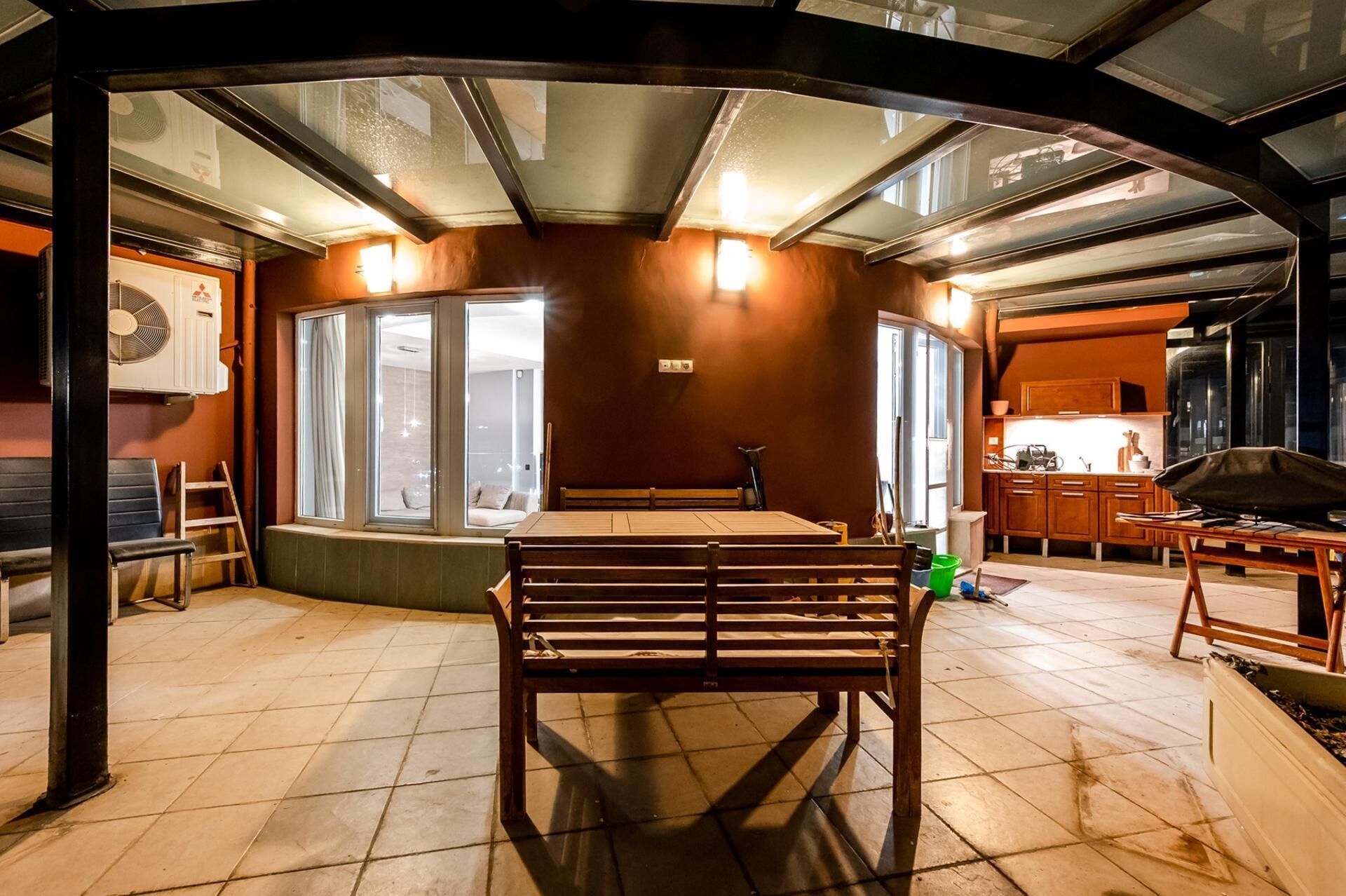Een eethoek met een tafel en banken, een keuken met houten kasten, en een groot raam dat natuurlijk licht binnenlaat.