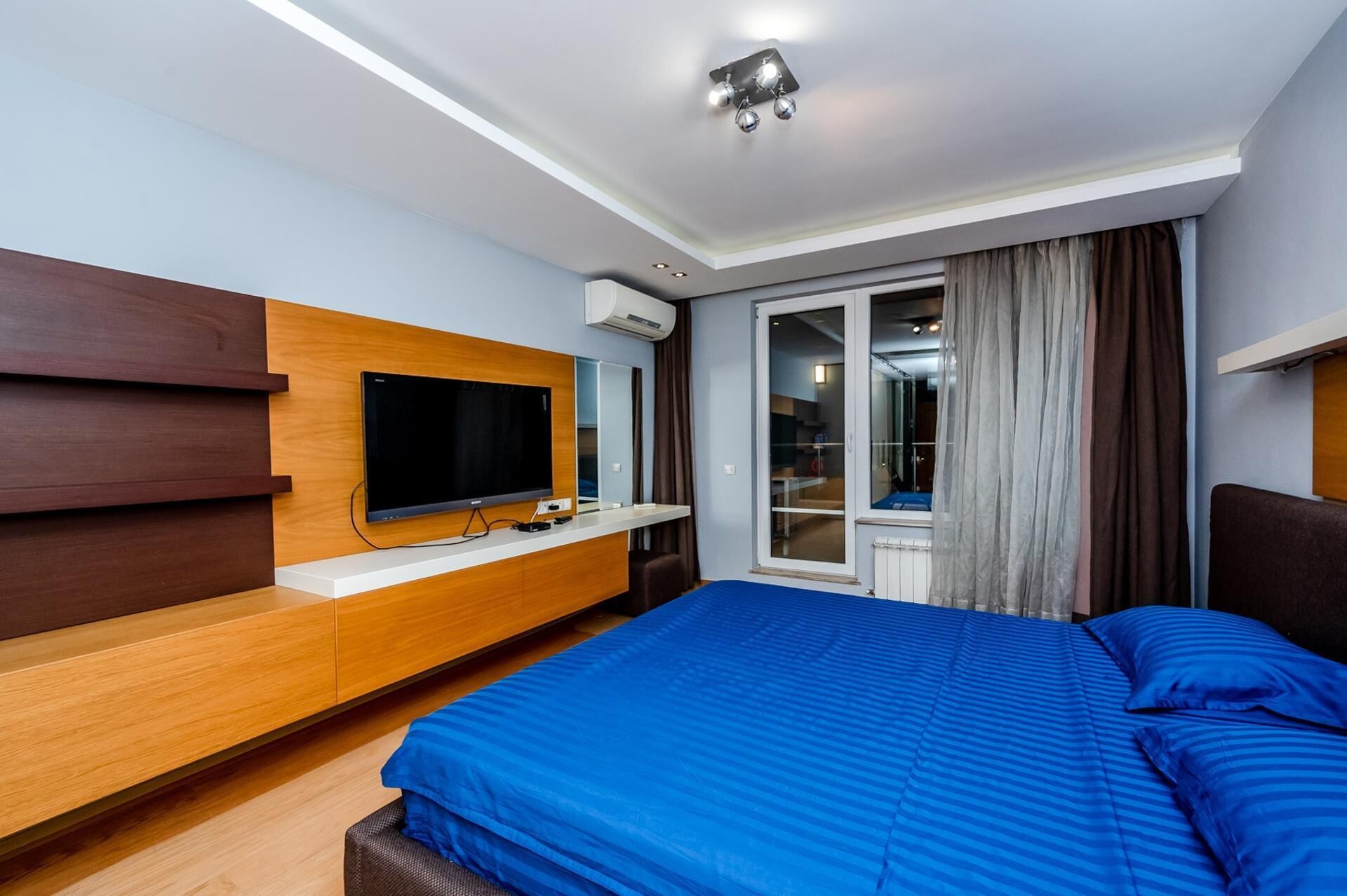 A modern hotel room with a large bed, a flat-screen TV mounted on a wooden console, a sliding glass door to the balcony, and a ceiling light fixture.