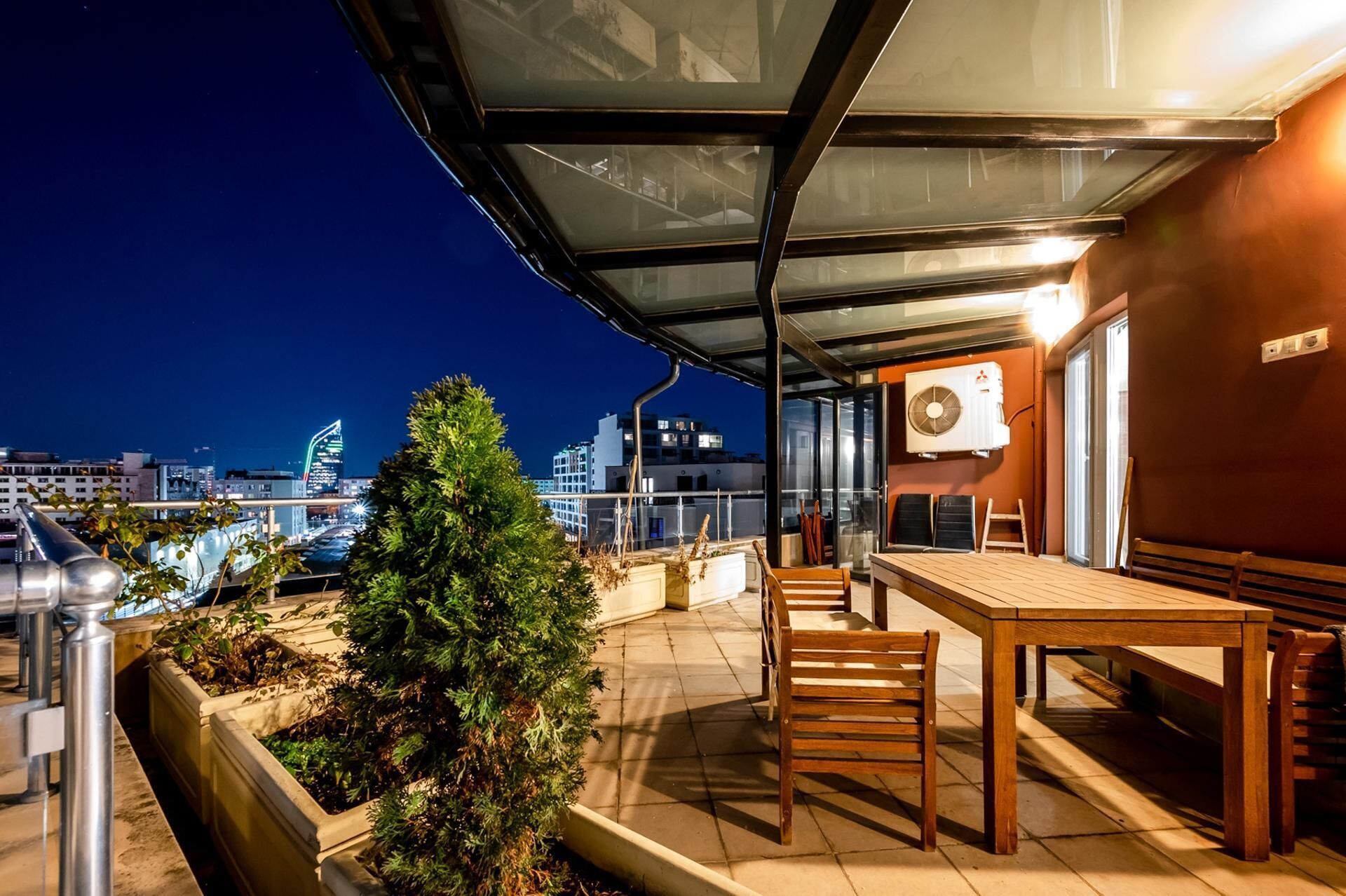 Une terrasse extérieure avec une table et des chaises, un petit arbre et une vue sur les toits de la ville illuminés la nuit.