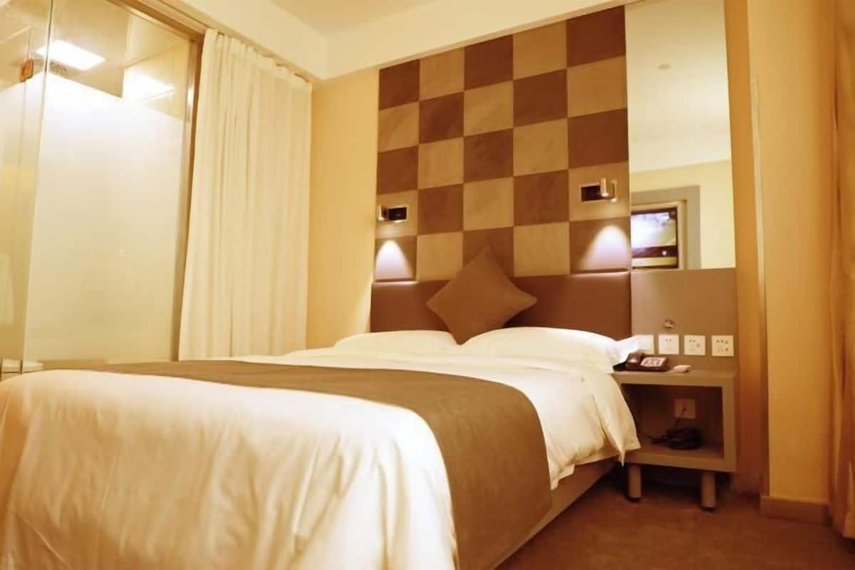 Une chambre d’hôtel avec une tête de lit à carreaux, un lit recouvert de draps blancs, une table de chevet avec une lampe et un téléviseur fixé au mur.