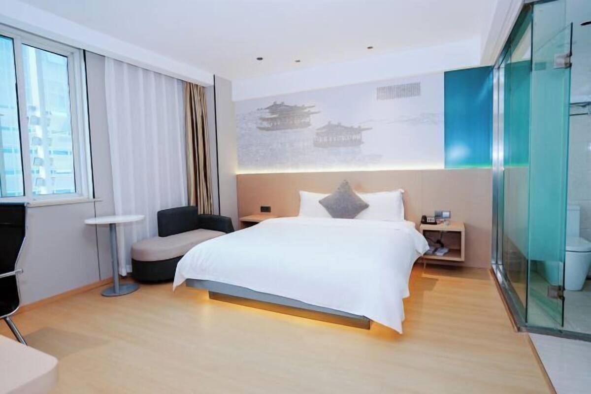 Ein modernes Hotelzimmer mit einem großen Bett, einer kleinen Sitzecke, einem Schreibtisch und einer Glaskabine, die das Zimmer vom Badezimmer trennt.