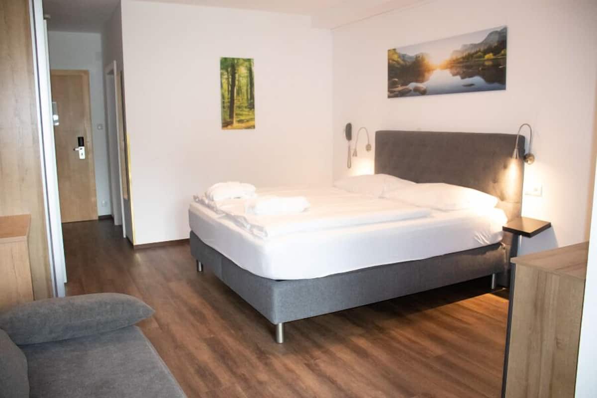 A bedroom with a bed, a bedside table, a nightstand, a wall-mounted lamp, and a painting.