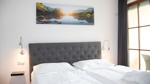 Un dormitorio con una cama grande, cabecera, dos lámparas de noche, un cuadro colgado en la pared que muestra un paisaje y una ventana con cortinas.