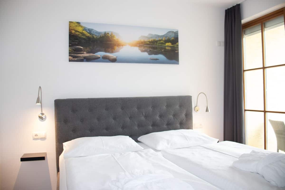 A bedroom with a large bed, a headboard, two bedside lamps, a wall-mounted picture of a landscape, and a window with curtains.