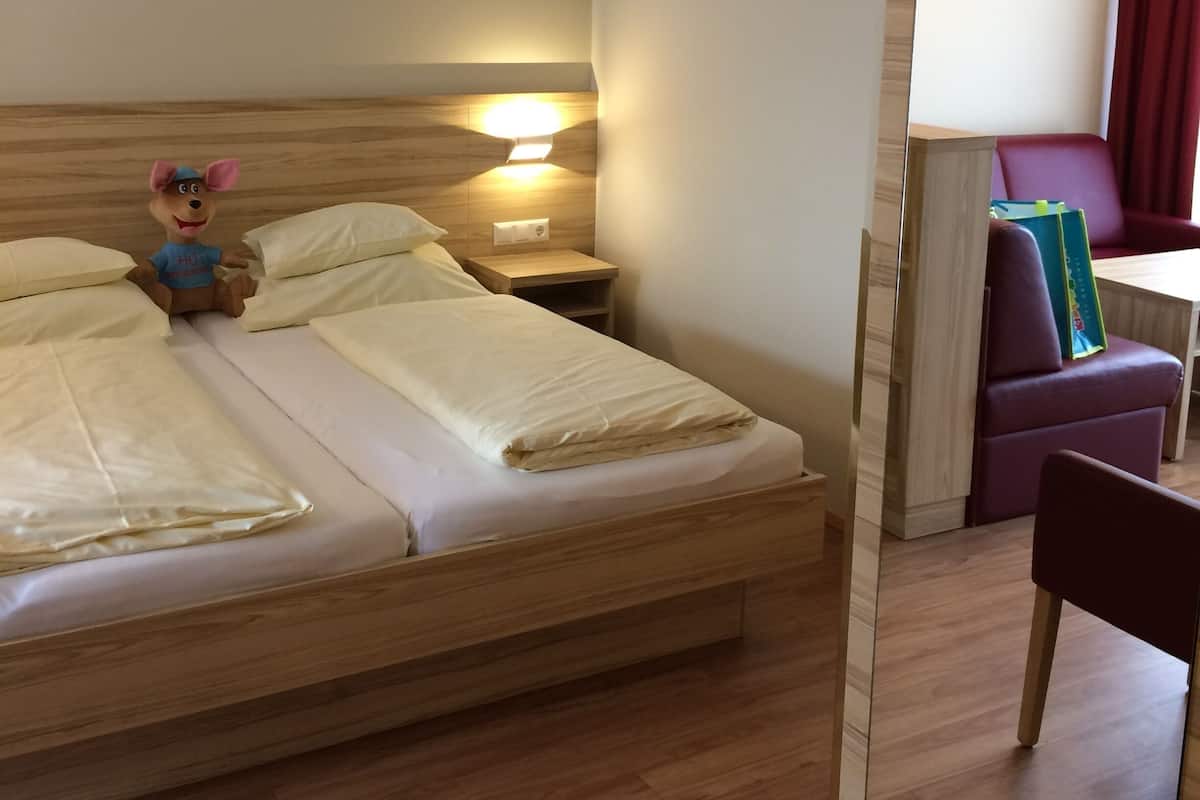 A hotel room with a wooden bed, a nightstand with a lamp, a plush toy on the bed, and a red chair in the corner.