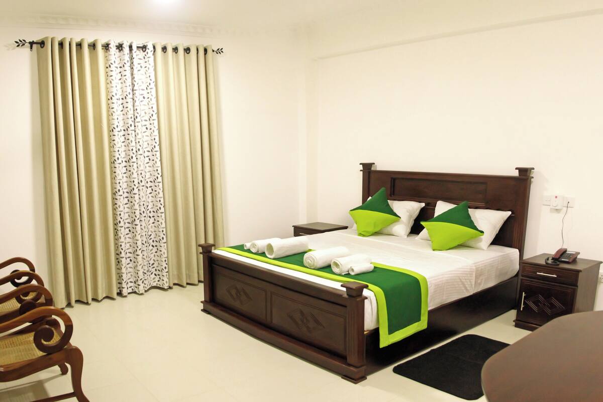 A neatly made bed with green and white bedding, a wooden nightstand with a telephone, and a chair with a woven seat.