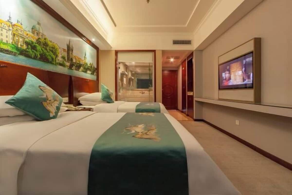 Habitación de hotel con dos camas, un espejo grande, un televisor y un cuadro de un paisaje urbano.