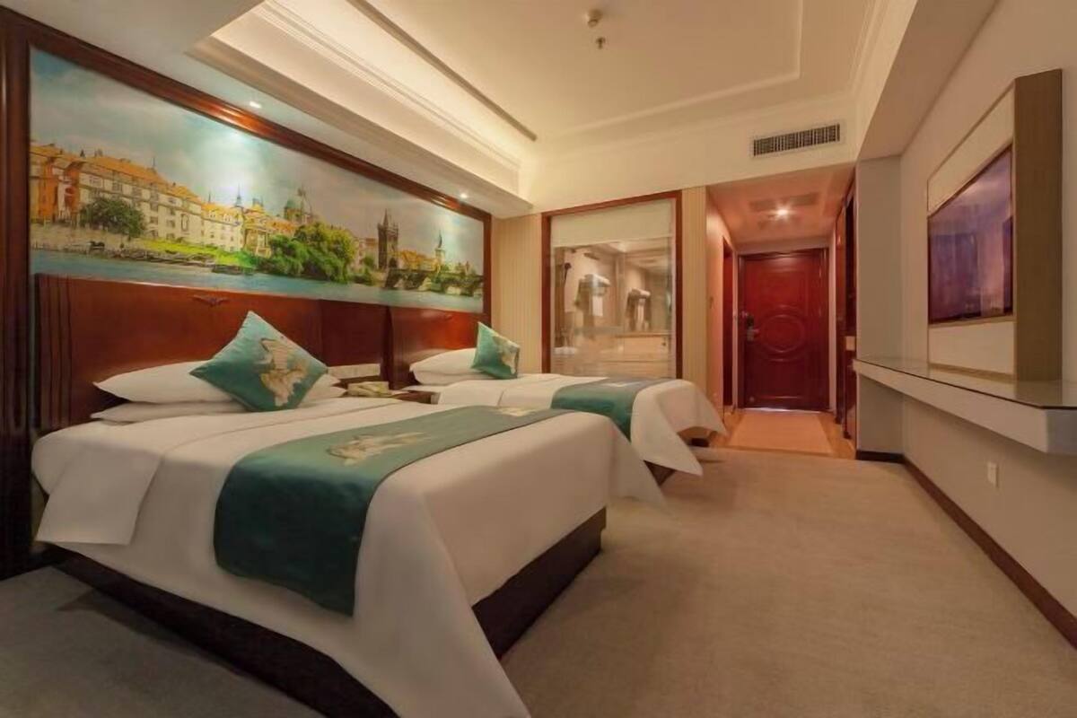 Habitación de hotel con dos camas, un cuadro de un paisaje urbano, un televisor y una puerta que da a otra habitación.