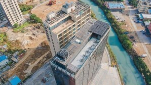 A high-rise building with multiple floors, a river running alongside, and construction sites nearby.