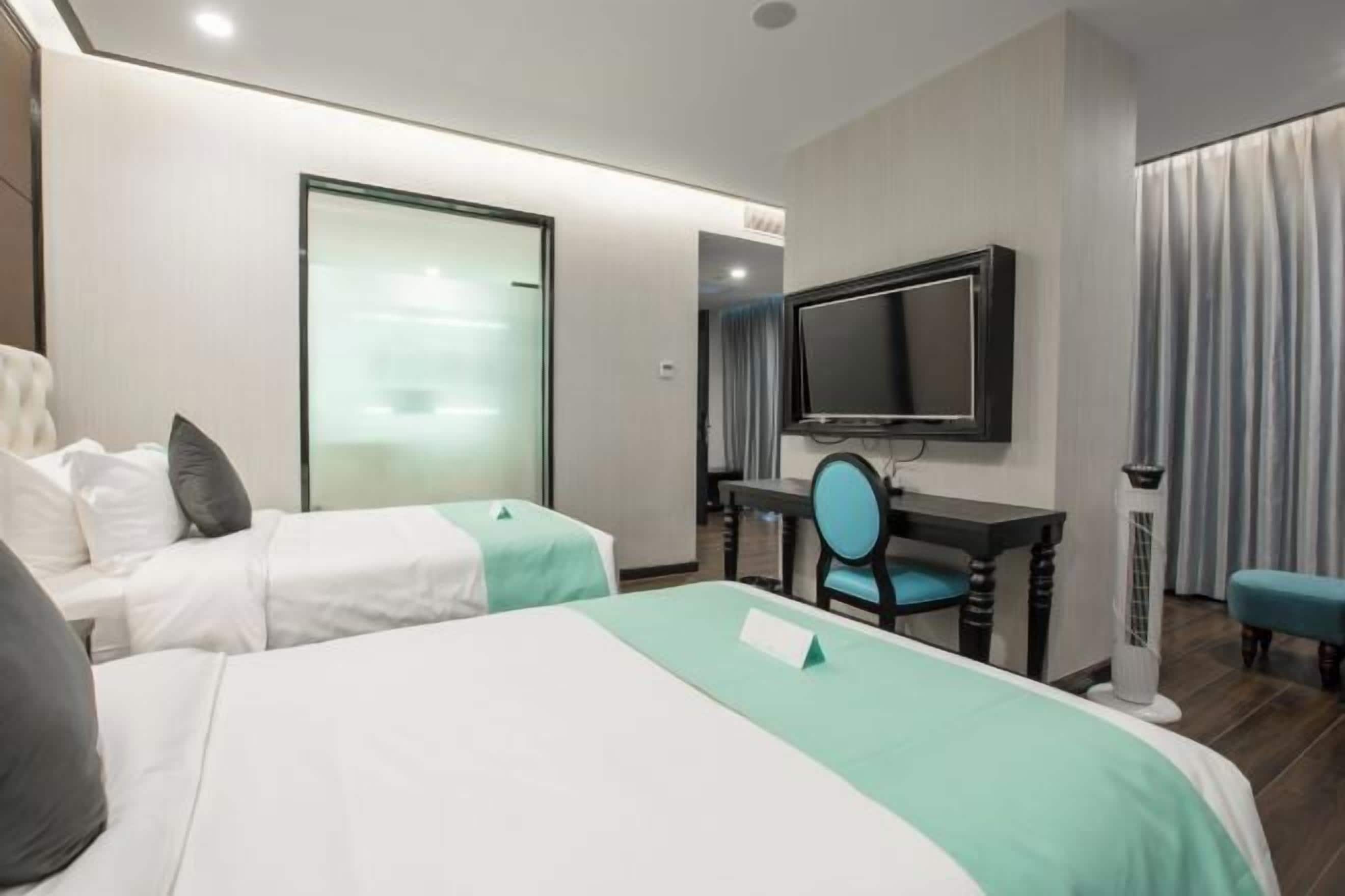 Quarto de hotel com duas camas, uma escrivaninha com cadeira, televisão e janela com cortinas.