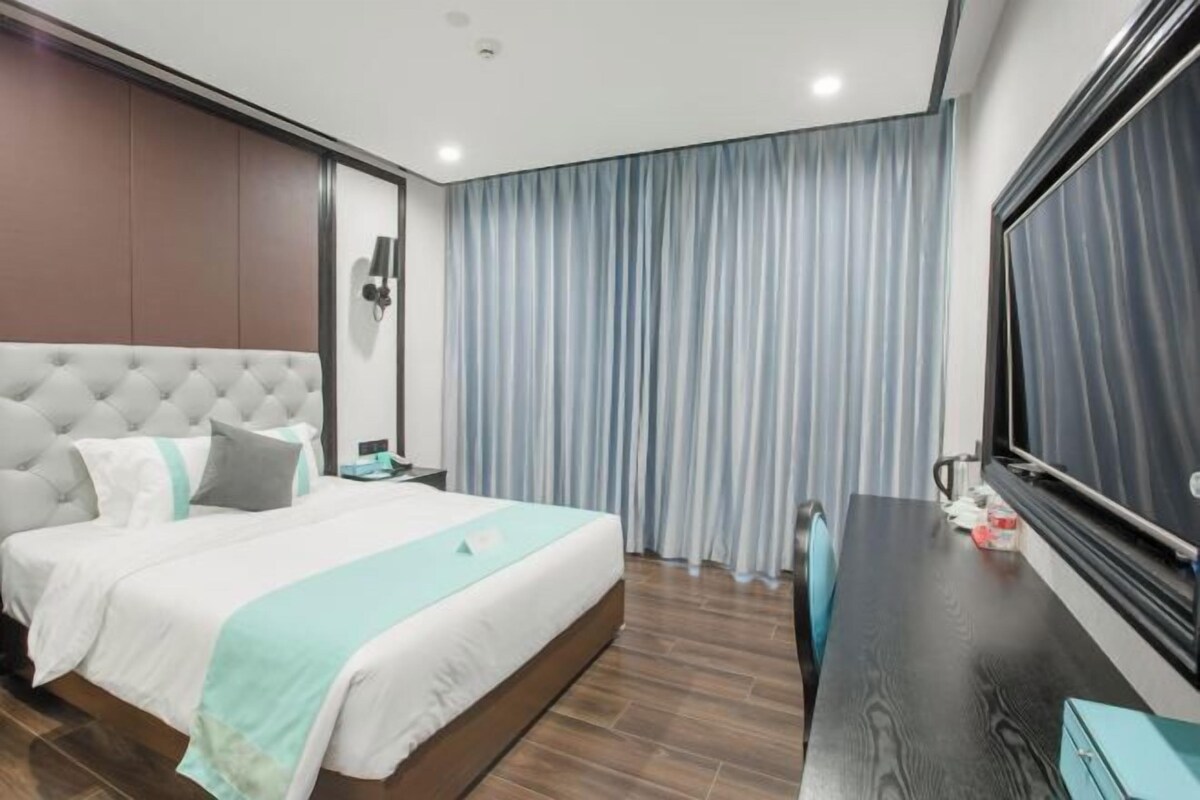 A modern hotel room with a large bed, a desk, a TV, and a window with curtains.
