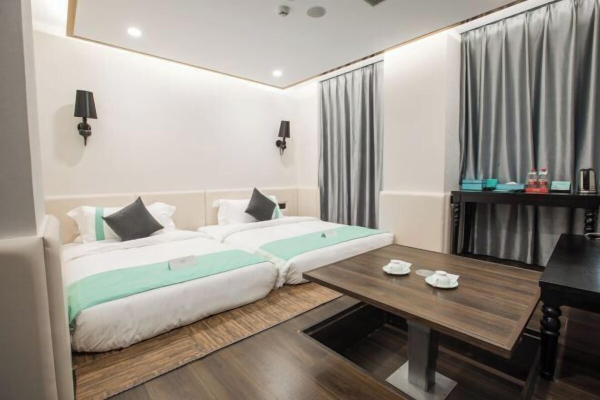 A hotel room with two beds, a wooden table, a small table with teapots, and a wall-mounted lamp.