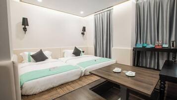 Quarto de hotel com duas camas, uma mesa de madeira, uma mesinha com bules e um abajur fixado na parede.