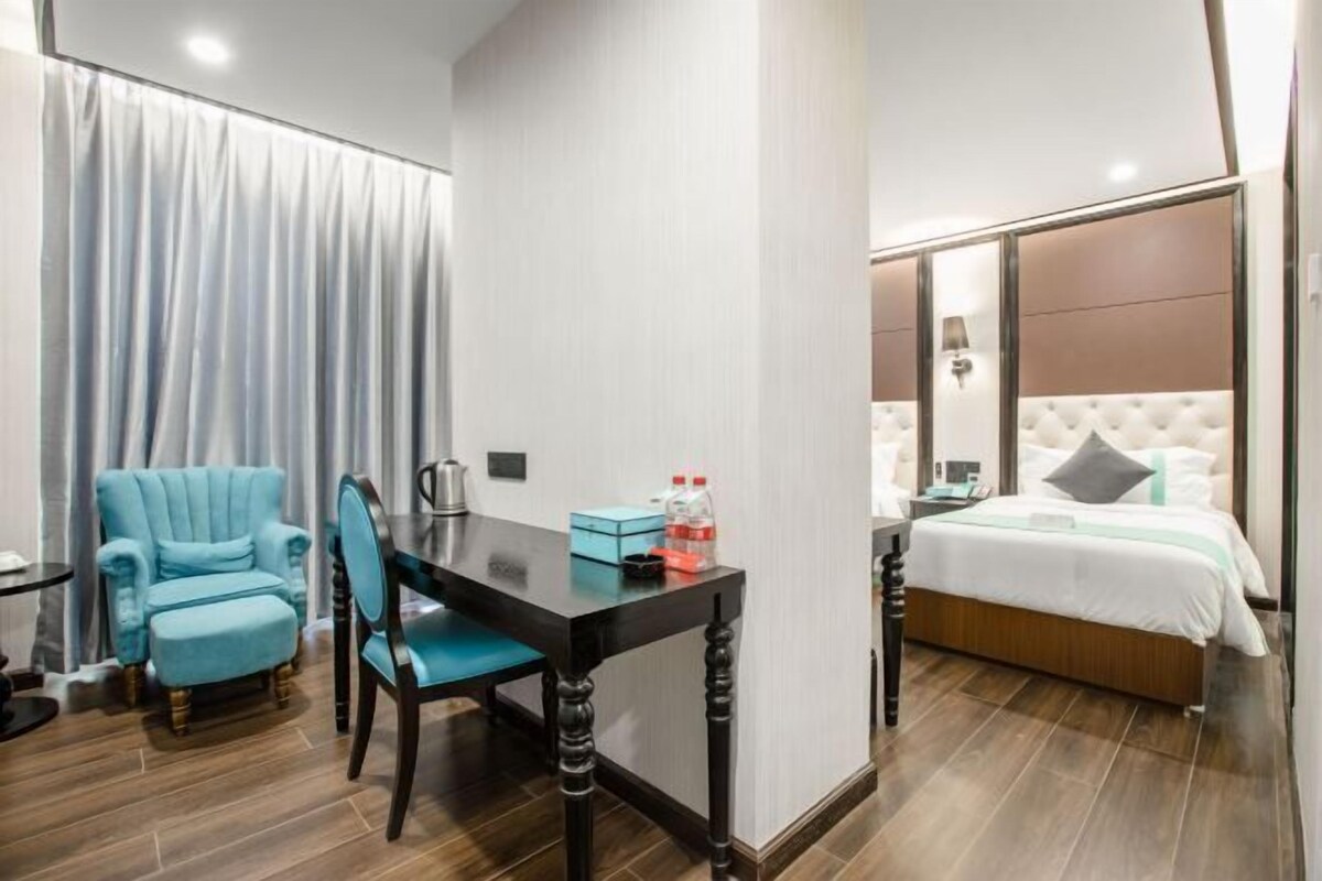 A hotel room with a bed, a desk with a chair, a blue armchair, and a window with curtains.