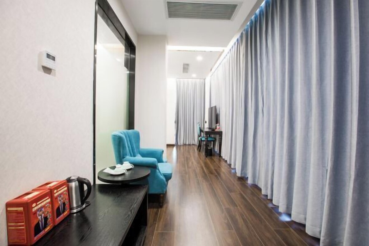 A hotel room with a wooden floor, a blue armchair, a small table with a kettle, and a TV on a stand.