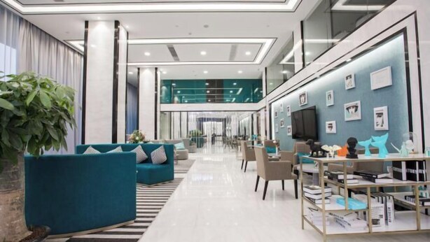 A modern hotel lobby with teal sofas, a coffee table, a bookshelf, and a large potted plant.
