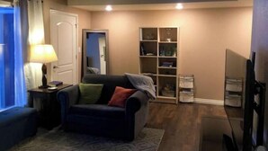 A cozy living room with a dark-colored sofa, a small table with a lamp, a bookshelf, and a television. - Bootlegger Bungalow - Downtown Anchorage (Anchorage)