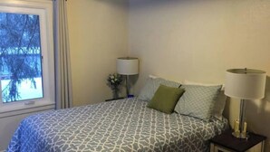 A bedroom with a patterned bedspread, a nightstand with a lamp, and a window with a view of trees. - Bootlegger Bungalow - Downtown Anchorage (Anchorage)