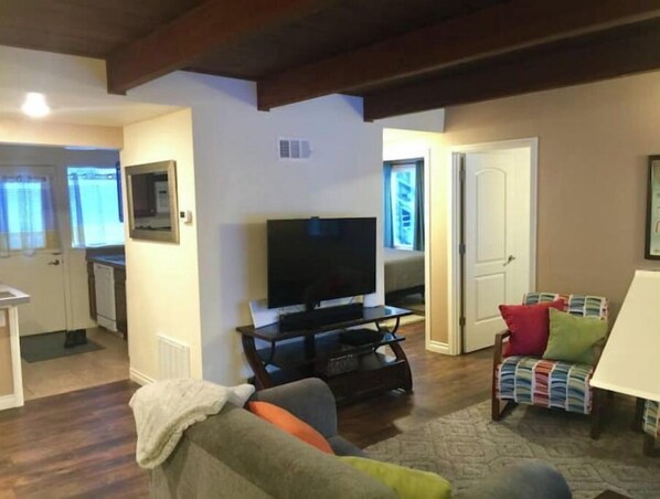 A living room with a television, a sofa, and a chair with cushions. There is a door leading to another room. - Bootlegger Bungalow - Downtown Anchorage (Anchorage)