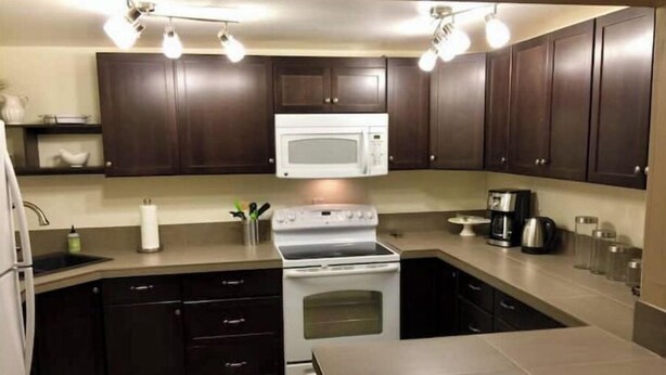 A modern kitchen with dark wooden cabinets, a white microwave, stainless steel appliances, and a large island. - Inlet View House - Downtown Anchorage (Anchorage)