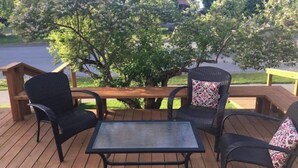 A wooden deck with two wicker chairs and a small table, surrounded by trees and a view of a road. - Inlet View House - Downtown Anchorage (Anchorage)