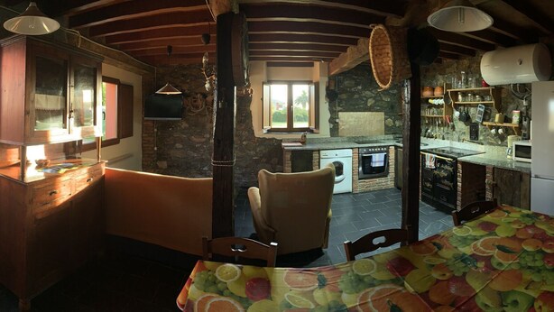 A rustic kitchen with stone walls, wooden beams, and a dining area with a fruit-patterned tablecloth. - EL PAJAR DE ONDINA (Cudillero)