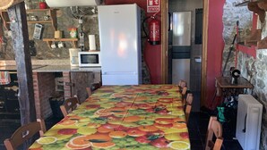 A rustic kitchen with a long dining table covered with a fruit-themed tablecloth, wooden chairs, a white refrigerator, and a fire extinguisher on the wall. - EL PAJAR DE ONDINA (Cudillero)