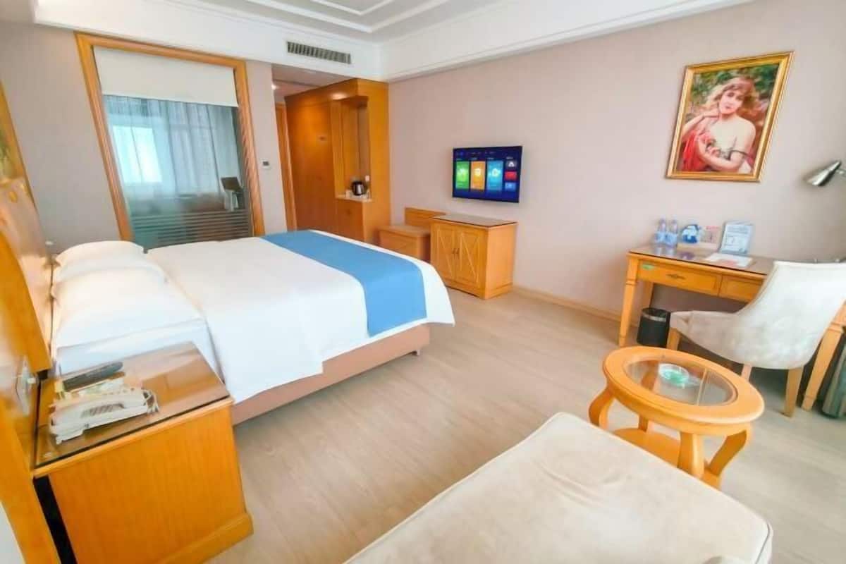 A hotel room with a bed, desk, chair, TV, and artwork on the wall.