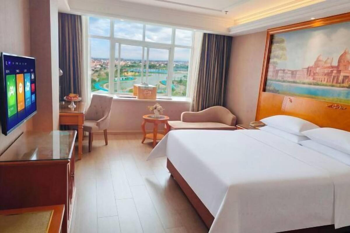 Habitación de hotel con una cama grande, un escritorio, una silla, una mesita, televisión y ventana con vistas a la ciudad.