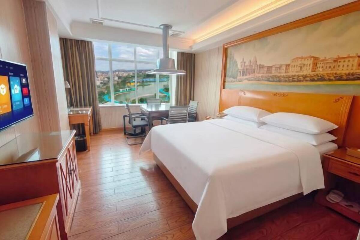 Habitación de hotel con cama, escritorio, televisor, ventana con vistas y un cuadro en la pared.
