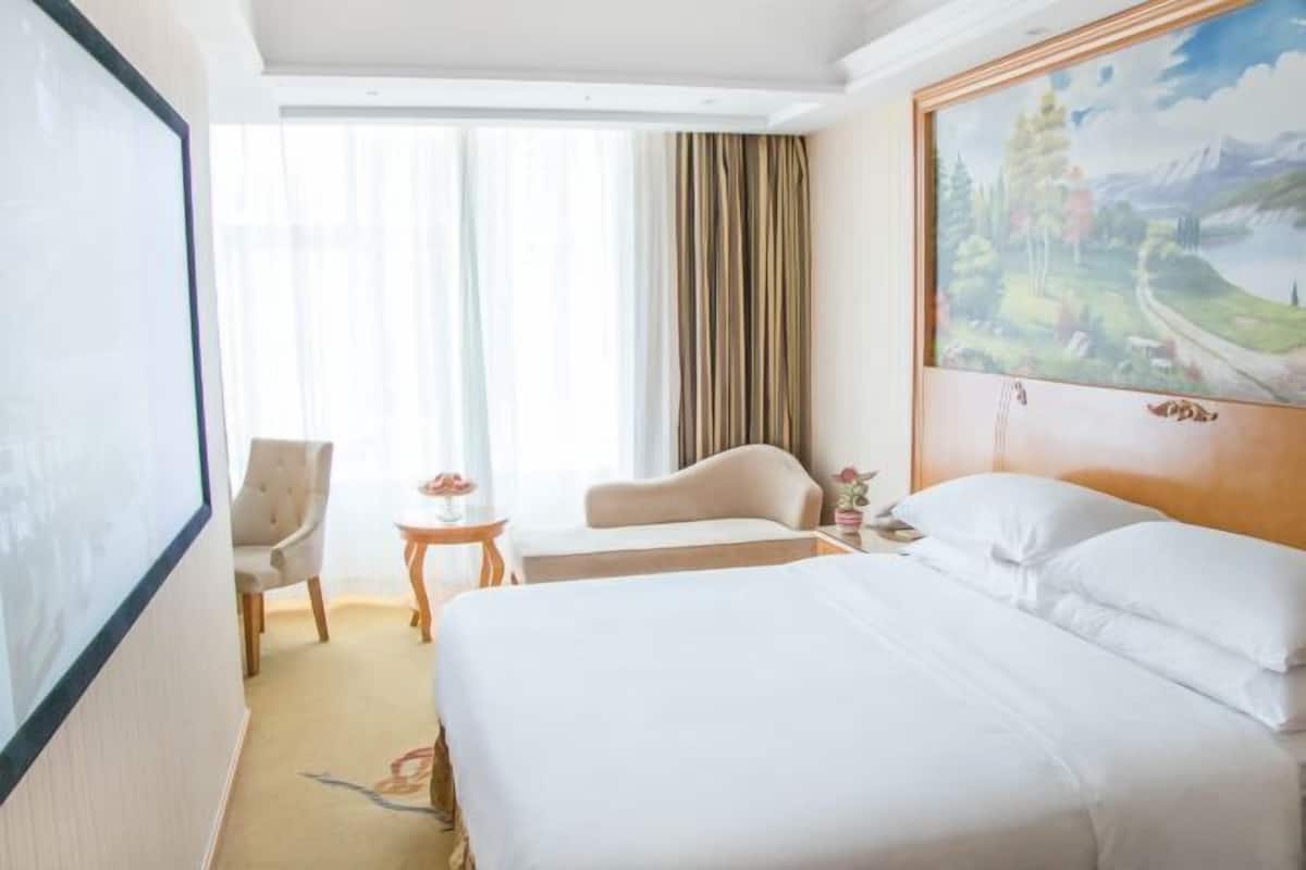 A hotel room with a large bed, a chair, a small table, and a painting on the wall.