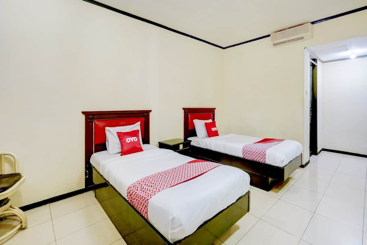 A hotel room with two beds, each with a red and white bedspread and matching pillows. There is a chair, a bedside table, and a door.