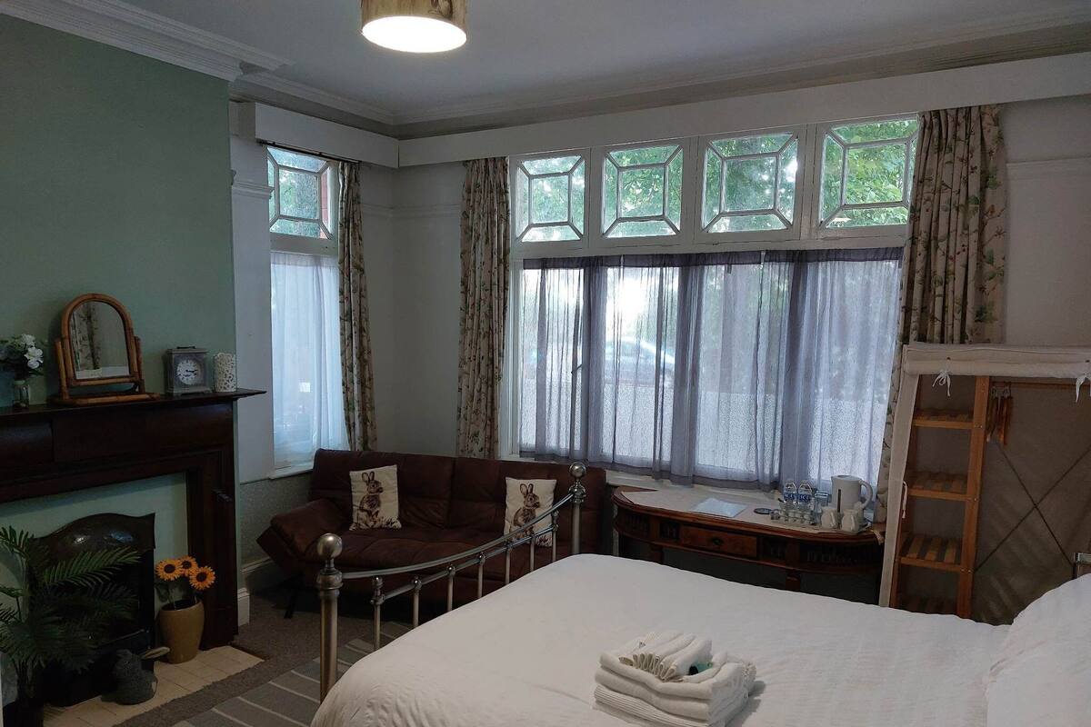 A hotel room with a bed, a sofa, a desk, a fireplace, and large windows.