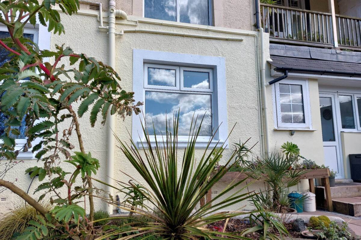 A residential building with a balcony, a window with a view of the sky, and various plants in the garden area.