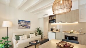 Uma sala de estar moderna com sofá, uma mesa de jantar com uma tigela de frutas, uma luminária e uma cozinha com micro-ondas e armários.
