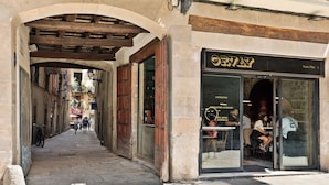 Uma rua estreita com um café chamado 'ORVAY' à direita, com uma porta de vidro e mesas ao ar livre visíveis pela janela.