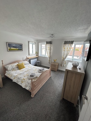 A bedroom with a wooden bed, a bedside table, a nightstand, a window with curtains, and a painting on the wall. - Immaculate 2-bed Cottage in Flamborough (Bridlington)
