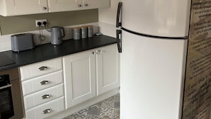 Eine Küche mit weißen Schränken, einer schwarzen Arbeitsplatte und einem weißen Kühlschrank. Auf der Arbeitsplatte befinden sich zwei Wasserkocher, ein Toaster und ein weiterer Wasserkocher. Der Boden ist mit einem gemusterten Fliesenbelag versehen.