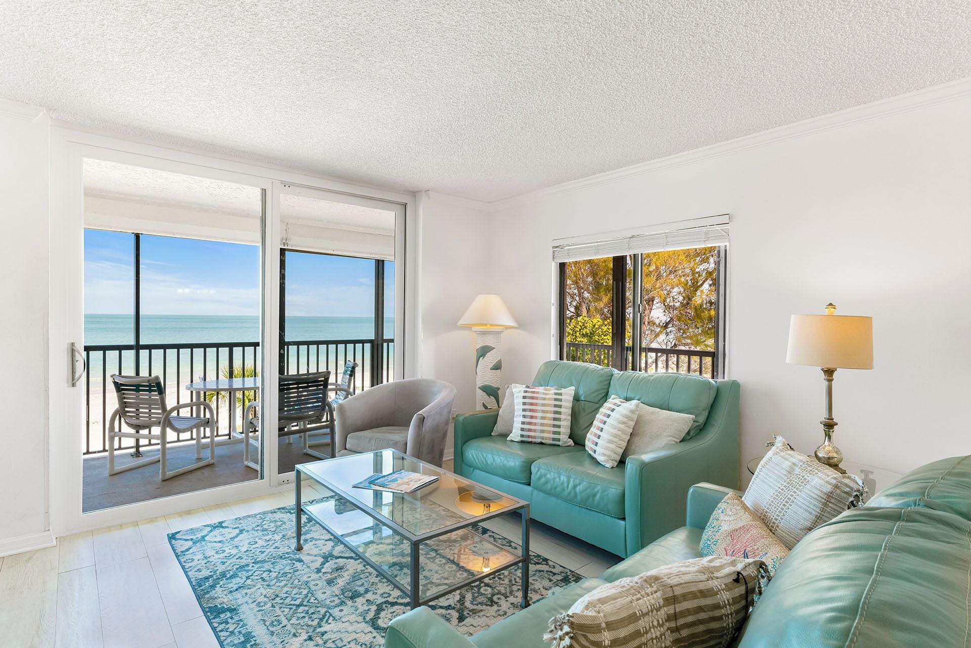 A living room with a teal sofa, a glass coffee table, and a view of the ocean through large windows.
