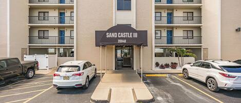 Un hotel de varios pisos con un cartel que dice 'SAND CASTLE III 20040' en la entrada. Hay espacios de estacionamiento frente al edificio.