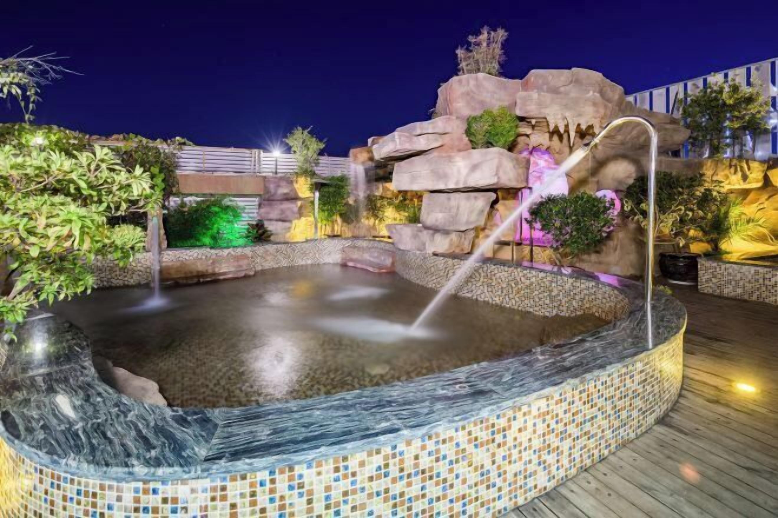 Ein Whirlpool mit Mosaikbordüre, umgeben von Steinen und Pflanzen, mit einem Wasserfall.