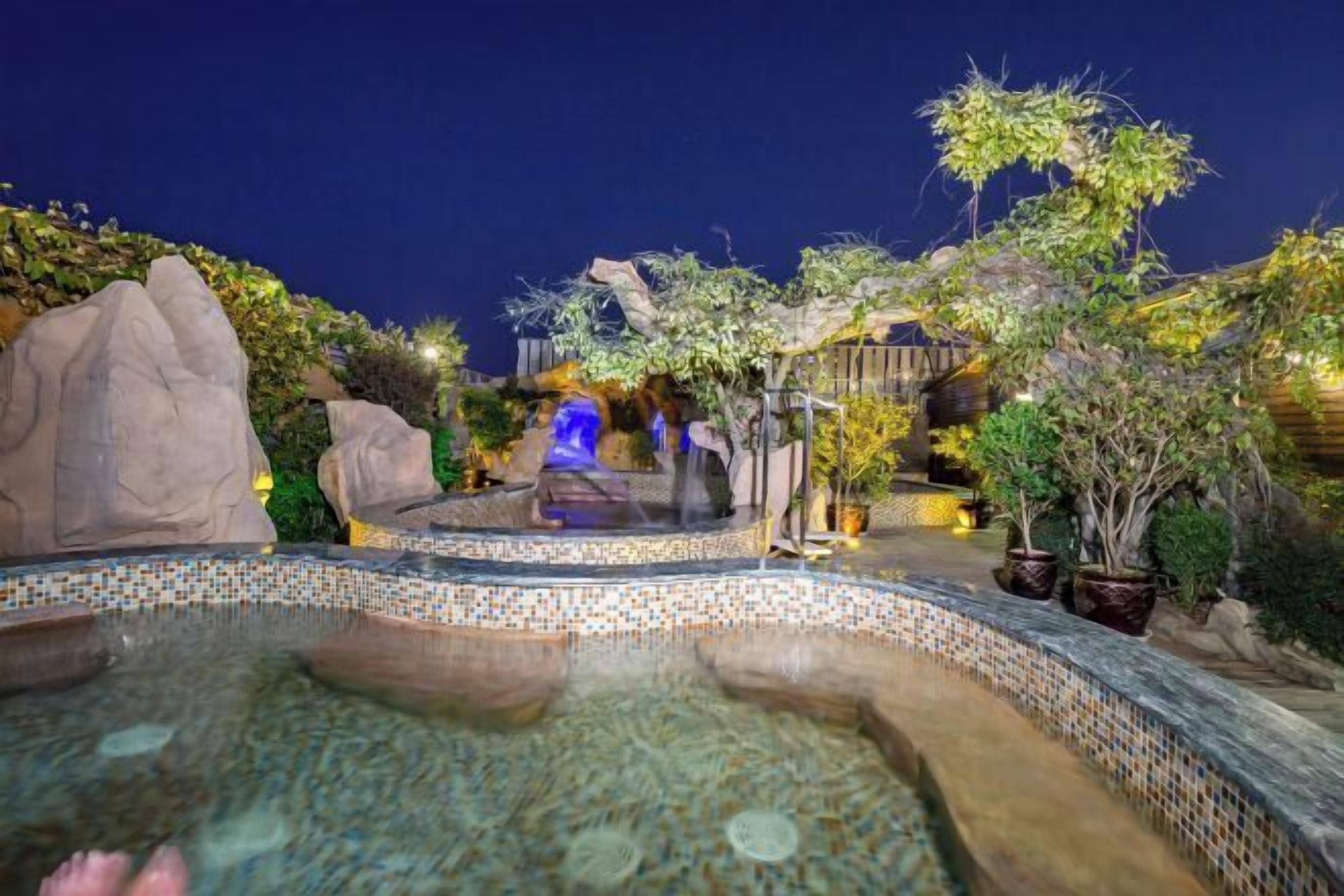 Zona de piscina exterior iluminada de noche, con un borde de mosaicos, rodeada de rocas y plantas en macetas.