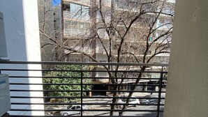 Un balcón con barandilla metálica, piso de baldosas y vistas a edificios y árboles.