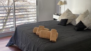 Un dormitorio con una cama grande, una mesita de noche con lámpara y vistas a árboles por la ventana.