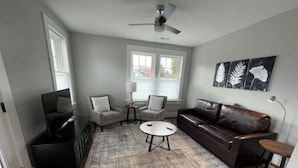 Un salon avec un canapé en cuir marron foncé, une table basse ronde et un tapis gris. On y trouve deux fauteuils, une petite table d’appoint et un ventilateur de plafond.