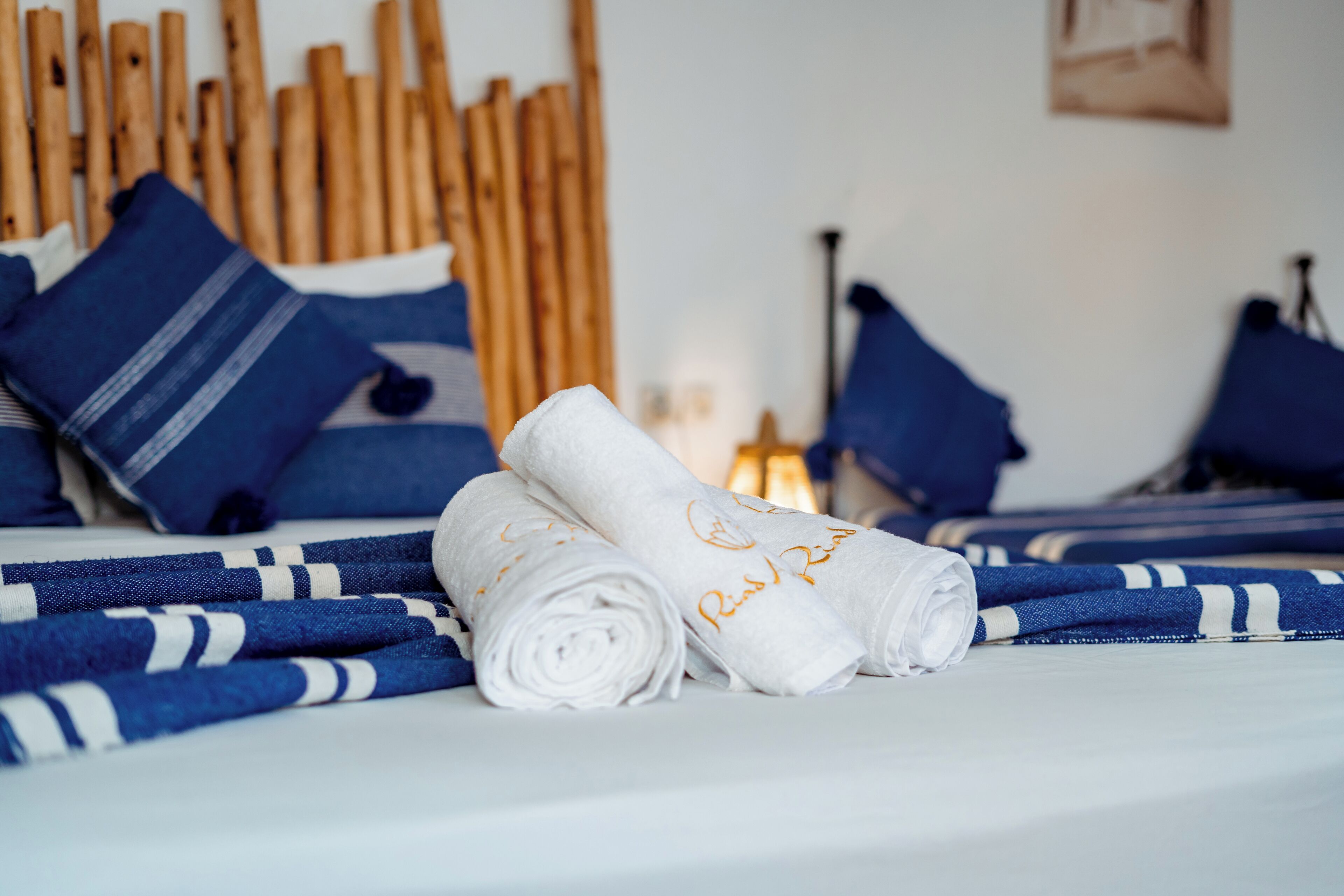 A neatly made bed with blue and white striped bedding, rolled white towels with embroidered text, and wooden headboards.