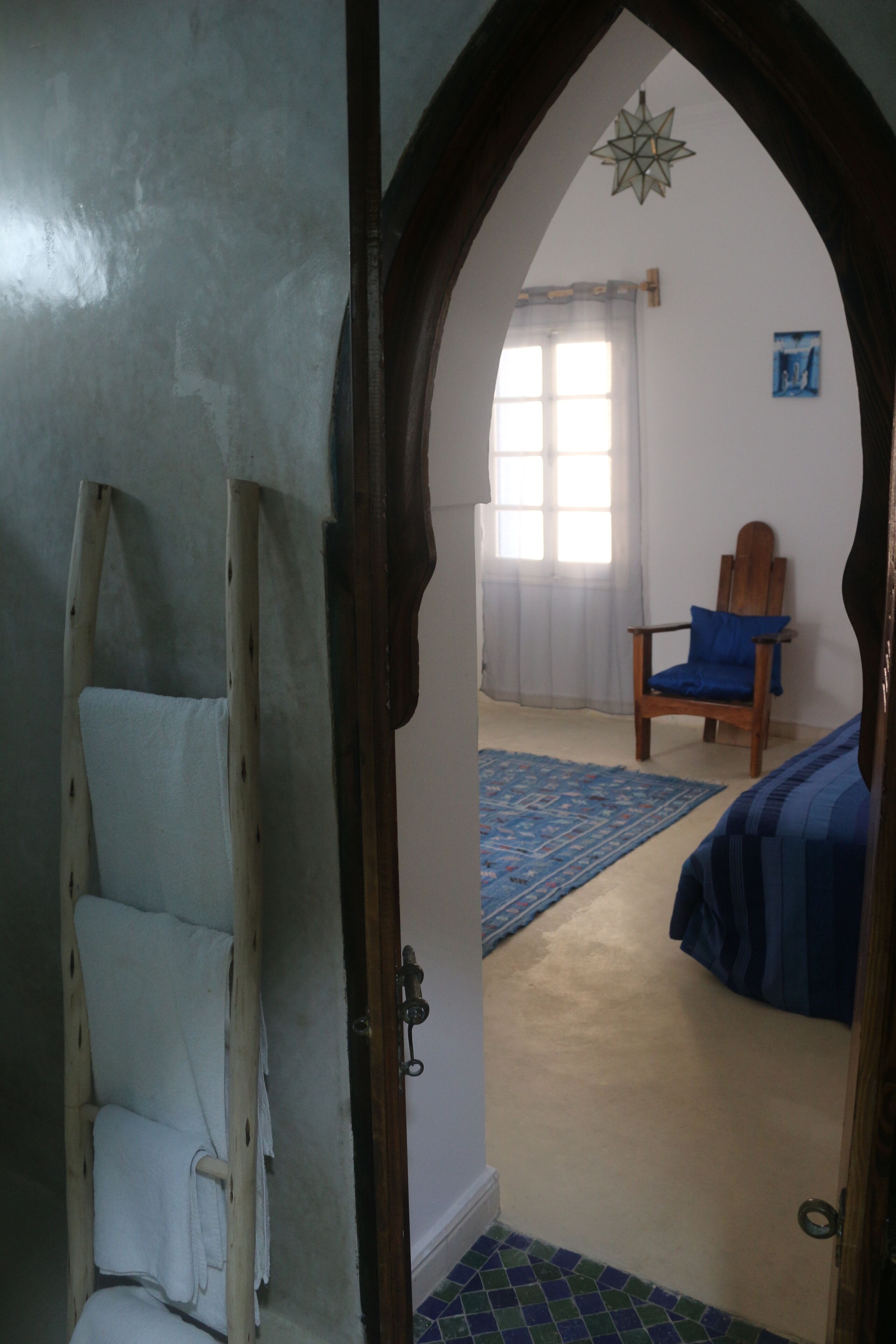 Une pièce avec une échelle en bois, un lit avec une literie bleue, une chaise avec un coussin bleu, un luminaire en forme d’étoile et une fenêtre avec des rideaux légers.