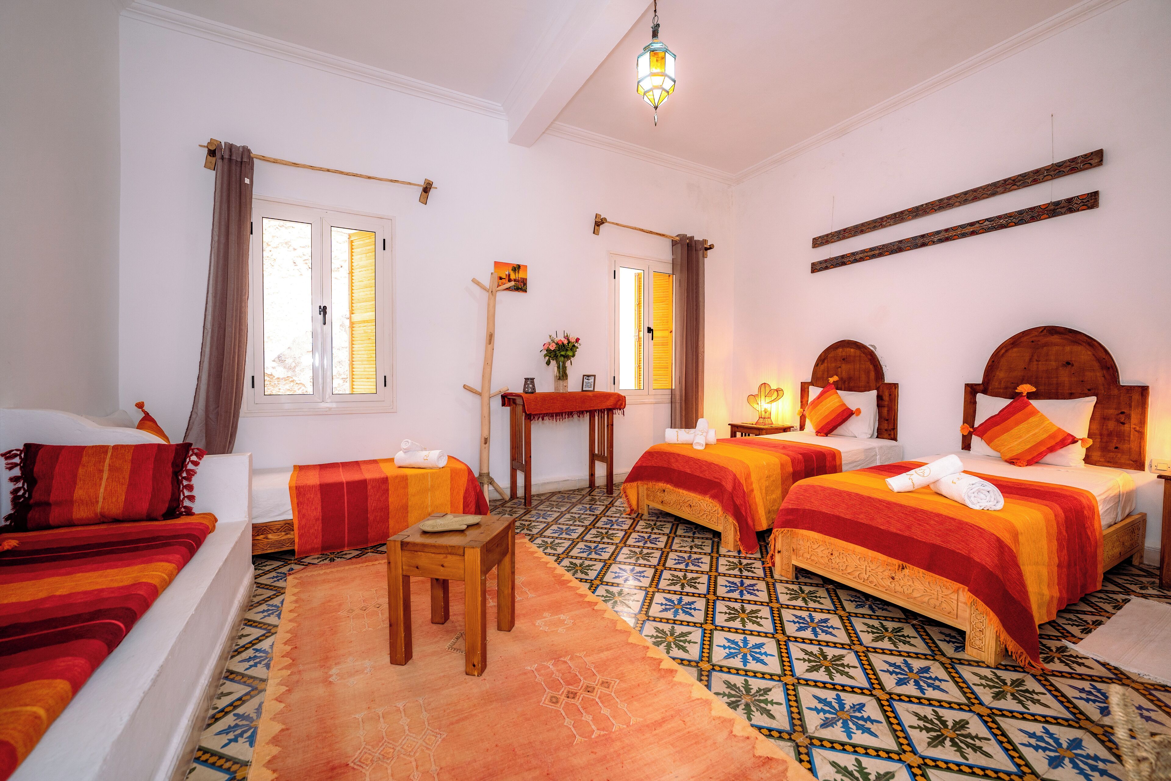 Habitación de hotel con dos camas, una mesita pequeña, una silla, una mesita de noche de madera, una lámpara de pie y un estante fijado a la pared.