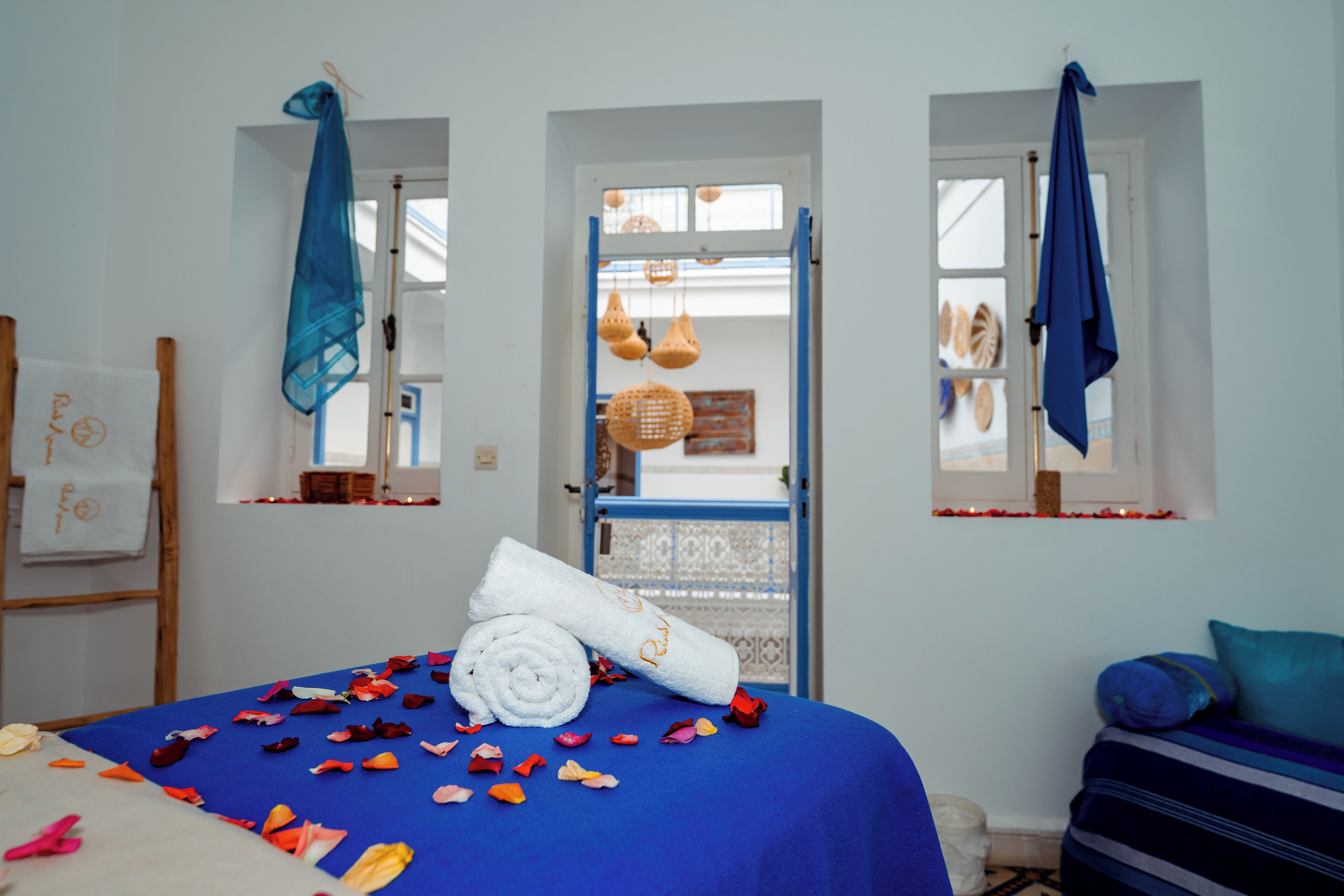 A room with a bed covered in a blue bedspread, a white towel, and scattered rose petals. There are two windows with blue curtains, a wooden ladder, and a blue and white patterned door.